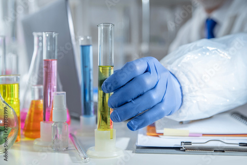 Chemistry laboratory scene with colorful chemical solutions in glass flasks and cylinders, scientist in gloves writing results, analytical experiment workspace.