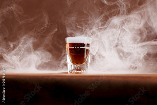 Whiskey in a traditional shot glass surrounded by a smokey bar top in a brownish, old western feel to the ambience. Intentionally shot with a warmer off camera lighting to emulate a traditional bar