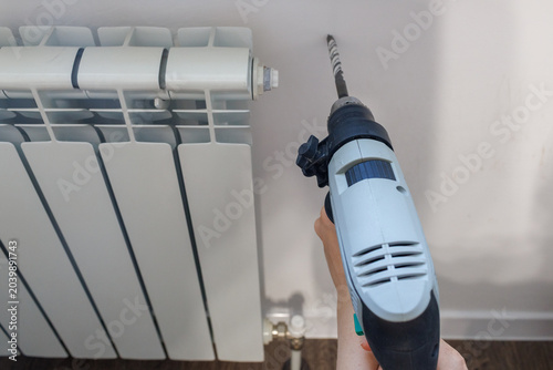 A first-person perspective of a hand using an electric drill on a white wall directly above a white heating radiator