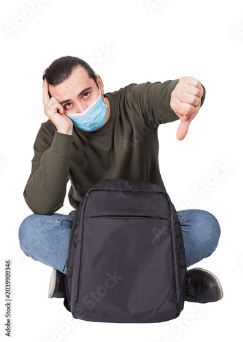 Картината върху платно Seated discontent man traveler with a backpack, wearing face mask as protection against viruses, shows thumb down gesture isolated on transparent background
