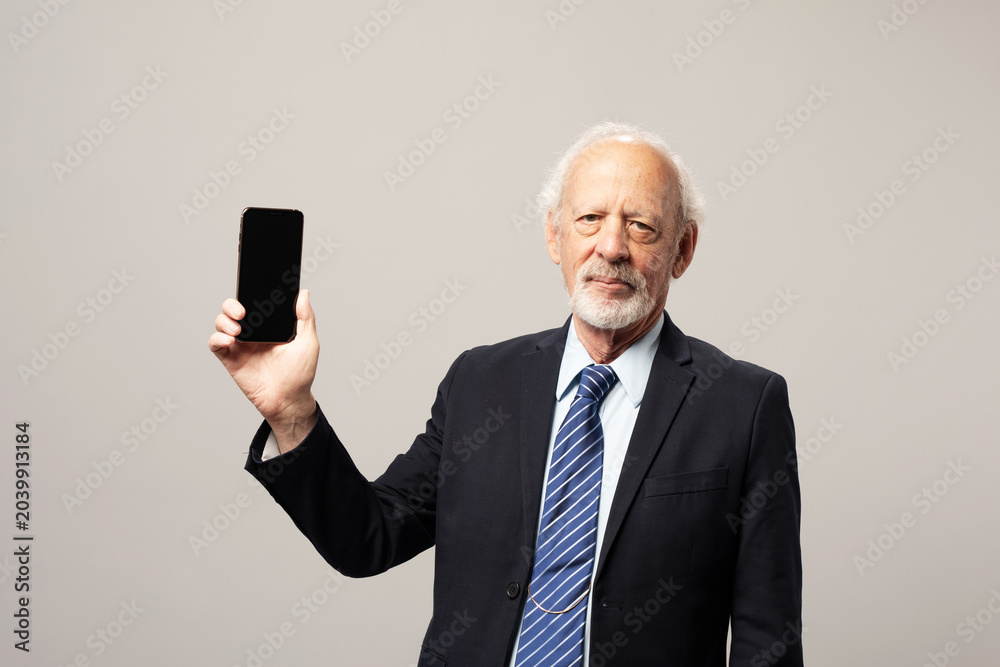 Naklejka premium Senior white man in a suit holding up a smartphone on a plain background