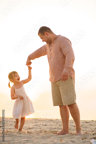 A father and his little daughter play and dance happily on a sandy lake beach at sunset. Happy childhood. Father's Day.
