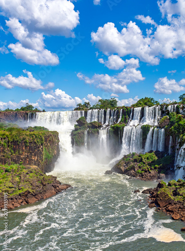 Breathtaking scenic view of the majestic Iguazu Falls with cascading water, lush green cliffs, and a vibrant blue sky with fluffy white clouds