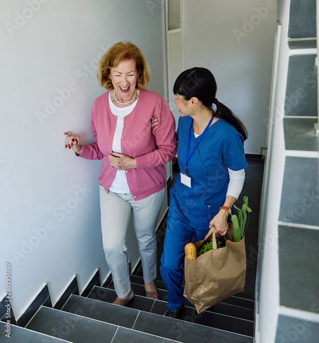 Doctor or nurse caregiver helping senior woman walking up the stairs and carrying groceries at home or nursing home