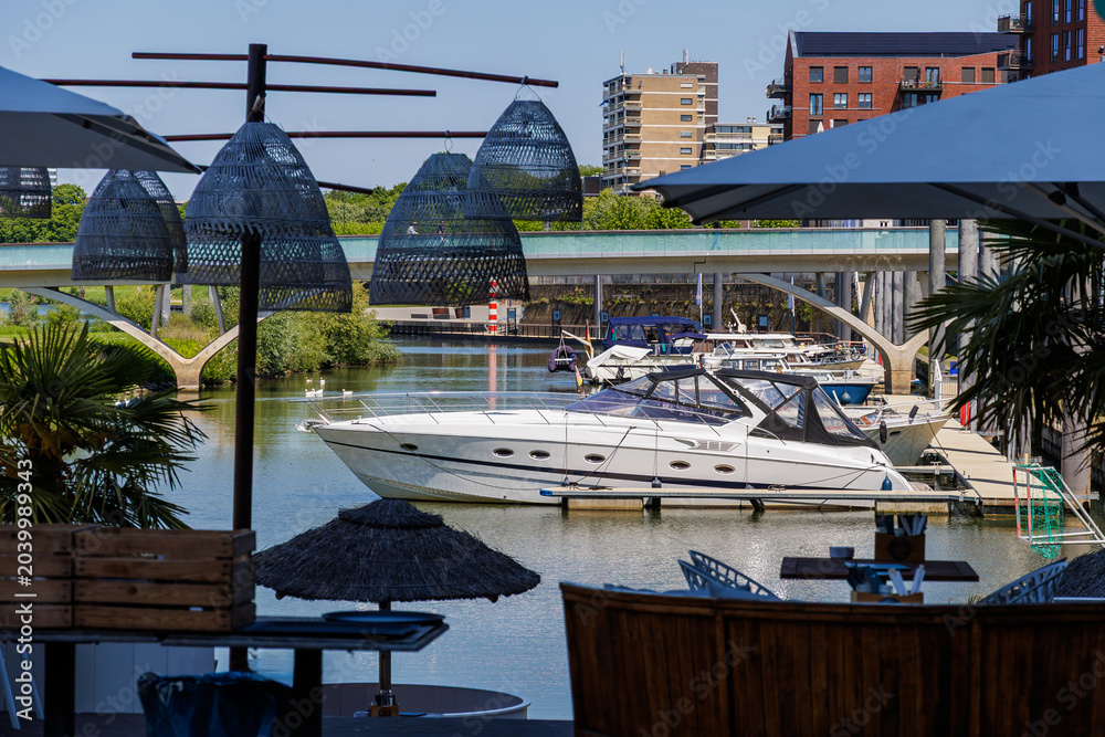 Naklejka premium Luxury yacht at marina viewed from waterfront terrace with umbrellas and city skyline.