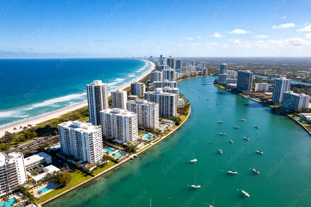 Obraz premium Aerial view of coastal city with high-rise buildings, turquoise waterway and sailboats under clear blue sky