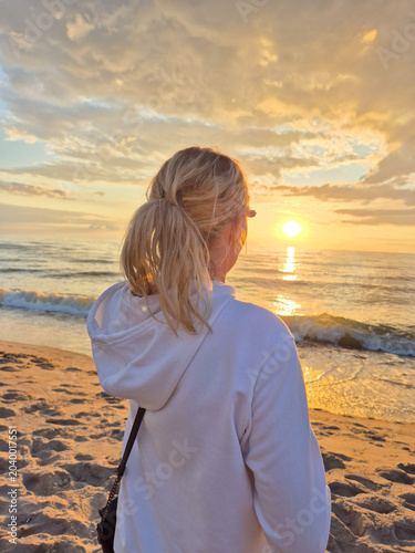 Blonde woman in white hoodie looking at a beautiful golden sunset over the sea.