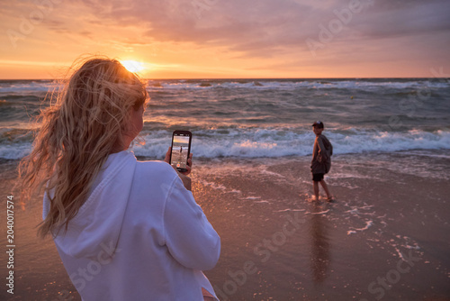 Proud mother holding smartphone and taking video of her son playing on the beach at sunset.