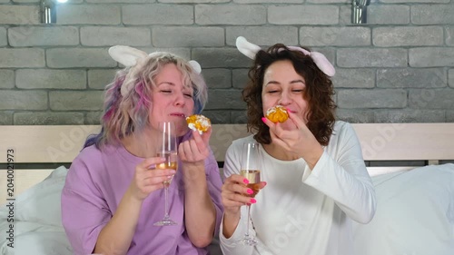 Two cheerful women friends eat cakes and drink champagne.
