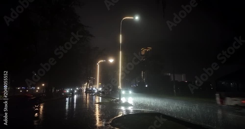 Heavy rain falls on a city street at night, where cars, motorbikes, and tuk-tuks drive under illuminated lamp posts, casting reflections on the soaked, shimmering road.