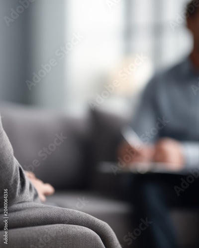 male patient sitting on sofa talking with psychologist during therapy session in cozy clinic office, mental health counseling concept, therapist listening and taking notes, soft natural light