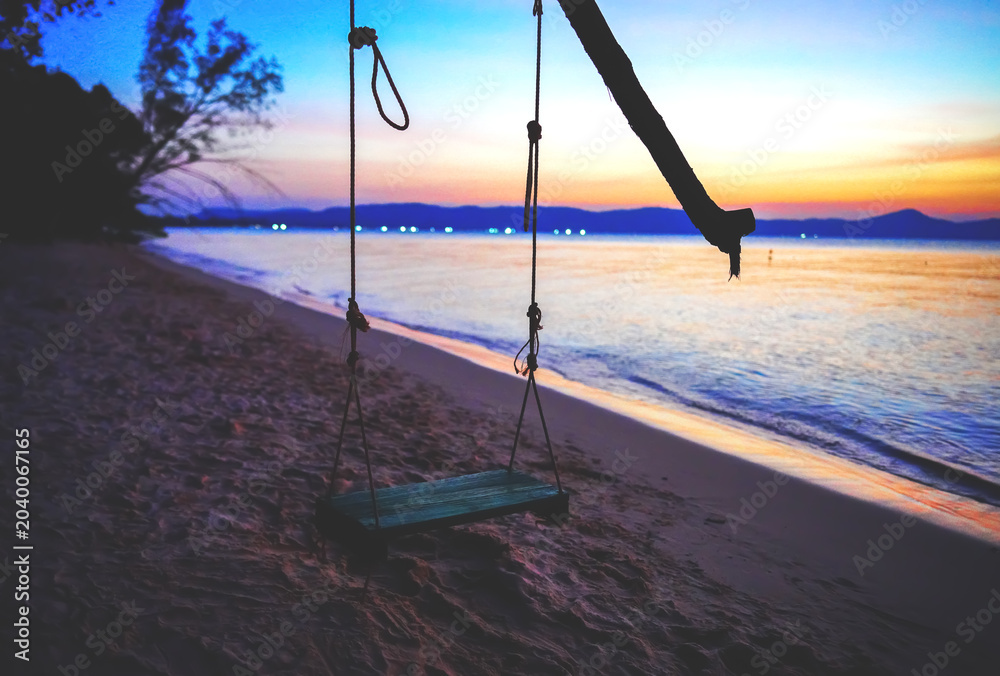 Fototapeta premium Swing on sea beach on sunset. Swing hangs on tree branch on shore tropical resort. Beach swing at ocean waves on sunset background. Vacation at sea with love on tropical paradise. 