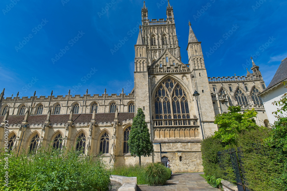 Obraz premium A historic stone cathedral with tall gothic windows and a prominent central tower stands bathed in sunlight under a vivid blue sky at Gloucester Cathedral - Gloucester - Gloucestershire - UK