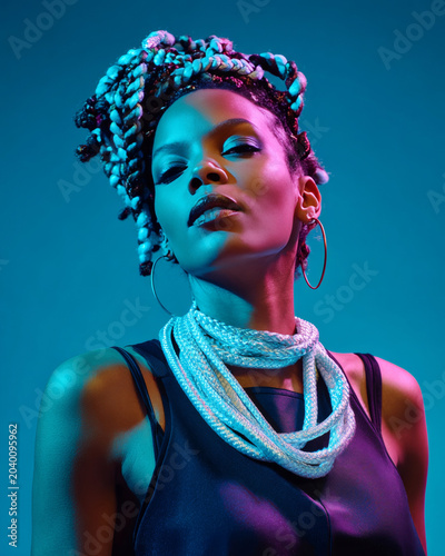 A woman with braided hair and hoop earrings is wearing a shiny necklace against a blue background