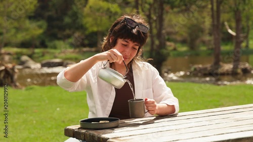 Woman pours tea from a teapot into a mug. She sits at a wooden picnic table. Grass and trees surround the area. River flows in the background. Her sunglasses rest on her head. She wears a white jacket