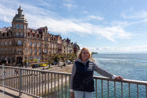 A happy senior woman stands on the Rhine Bridge with the historic architecture of Seestrasse and Lake Constance in the background