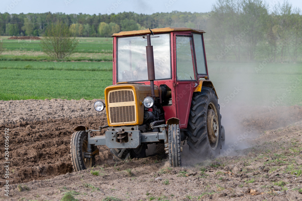Obraz premium Vintage agricultural tractor plowing dry soil on a farm field raising dust clouds during spring cultivation work in rural countryside, traditional farming machinery.