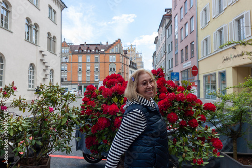 A happy senior woman with blonde hair and glasses posing by blooming red rhododendron bushes on a street in the historic Altstadt