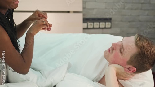Sensual multiethnic couple playfully eating grapes on the bed in the bedroom. A romantic guy and a girl are having a good time together, looking at each other, enjoying a tender moment. Happy loving