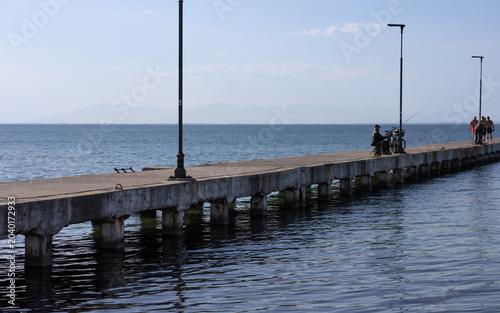 fisherman sitting on a concrete pier over the blue sea during a sunny day with distant mountains