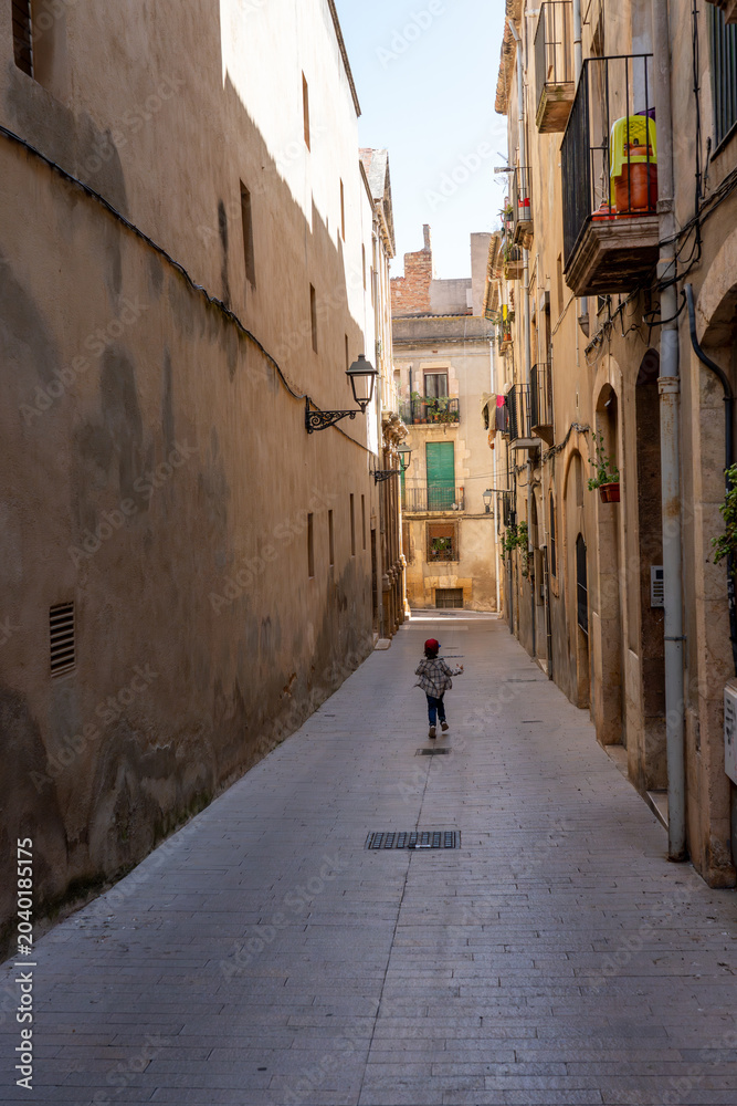 Obraz premium Back view of a small child walking down a quiet, sunlit cobblestone street lined with tall ancient buildings and traditional balconies in a historic Mediterranean town.