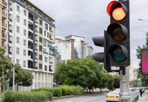 close-up shot of a city traffic light showing a red signal against a blurred urban background with modern apartment buildings, lush trees, and street traffic