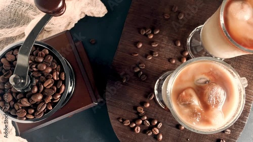 A flat lay shot reveals a vintage manual grinder filled with dark coffee beans alongside two glasses of iced coffee with milk, accented by scattered beans on a rustic wooden board.