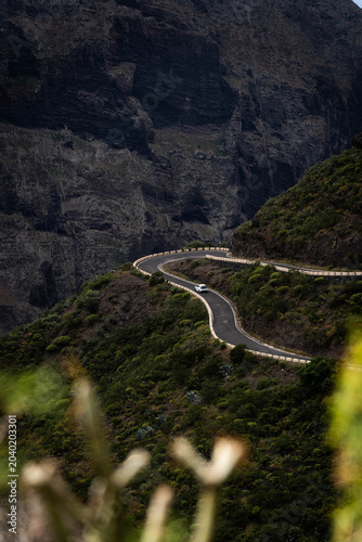 A solitary white car drives along a dramatic, winding serpentine mountain road through the deep volcanic canyons of Masca Valley in Tenerife.
