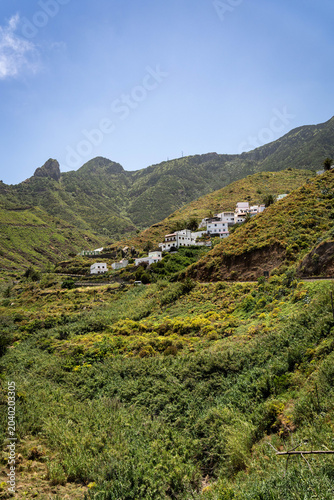 A picturesque view of traditional white houses built on the steep, lush green terraced hillsides of the Anaga mountains under a bright blue sky.