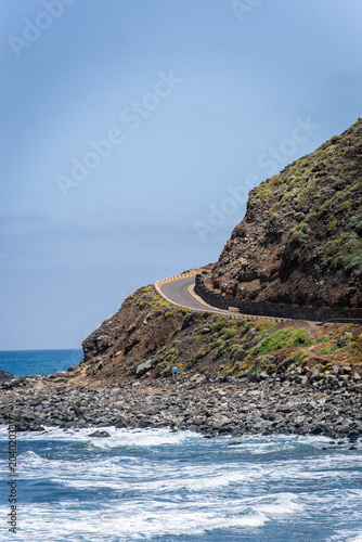 A beautiful asphalt coastal road wrapping around a volcanic hill next to crashing white ocean waves and a rocky beach on a clear blue day.