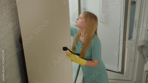 White house technician scrubbing tiled wall, blonde woman in mint uniform and yellow gloves inspecting grout and working upward, focused professional motion in bright bathroom setting, close side view
