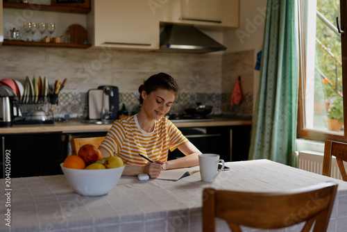 Quadro su tela A woman sits at a kitchen table writing in a notebook, bathed in natural light with a fruit bowl and coffee mug nearby, conveying a calm, homey morning mood