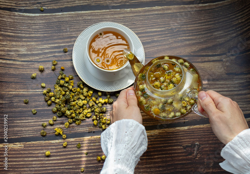 Photos Top view of female hands pouring freshly brewed chamomile and chrysanthemum tea from a glass teapot into a cup