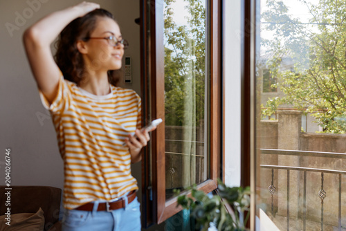 Photos A woman in striped shirt stands by an open window, basking in daylight, lost in thought