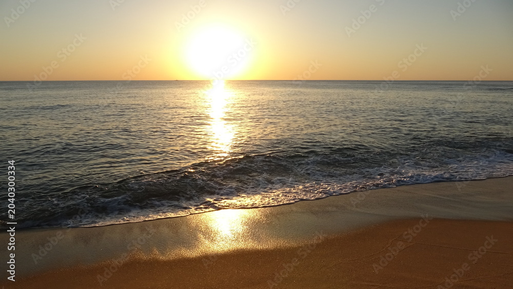 Obraz premium Majestic Sunrise over East Sea Horizon. The bright sun is rising over the ocean horizon, casting a strong golden reflection. Gentle waves with foam wash onto the sandy foreground beach.