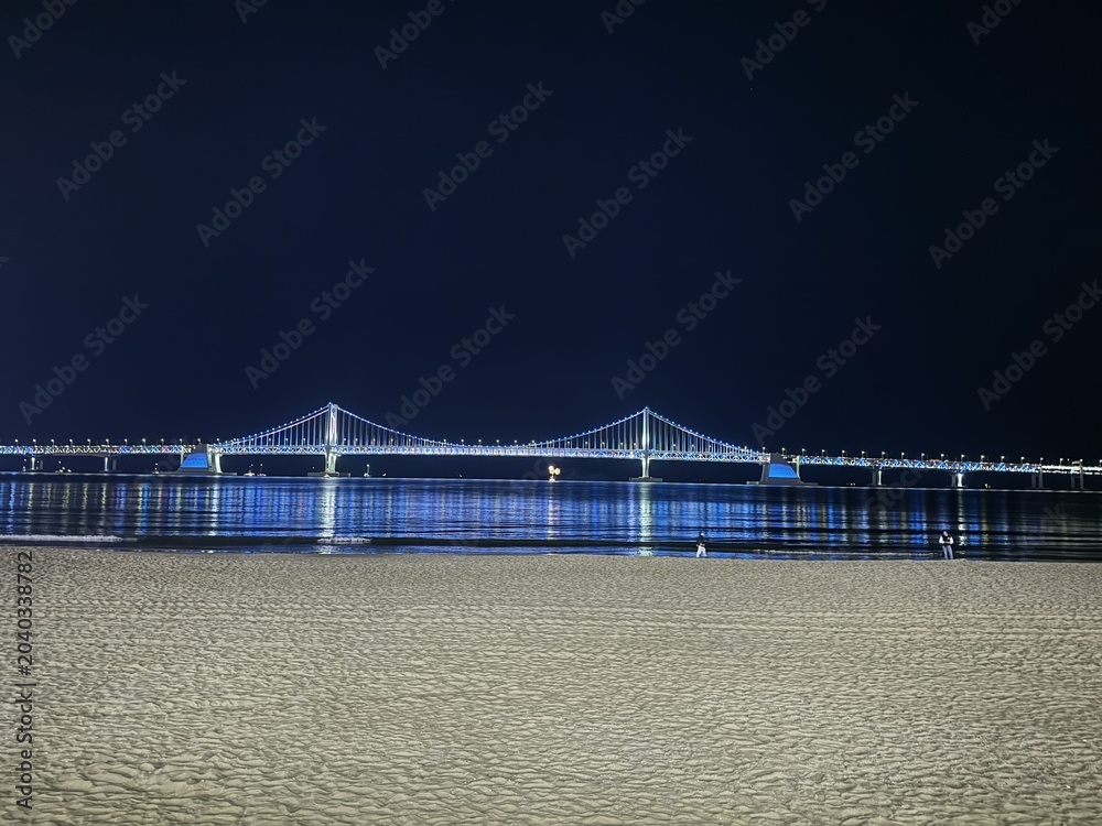 Fototapeta premium lluminated Suspension Bridge Glowing Blue at Night, Reflected on Calm Sea with Sandy Beach Foreground