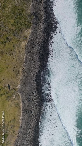 Aerial view of the coastline in Raglan, New Zealand, showing the waves crashing onto the rocky beach. The photo captures the beauty of the natural landscape.