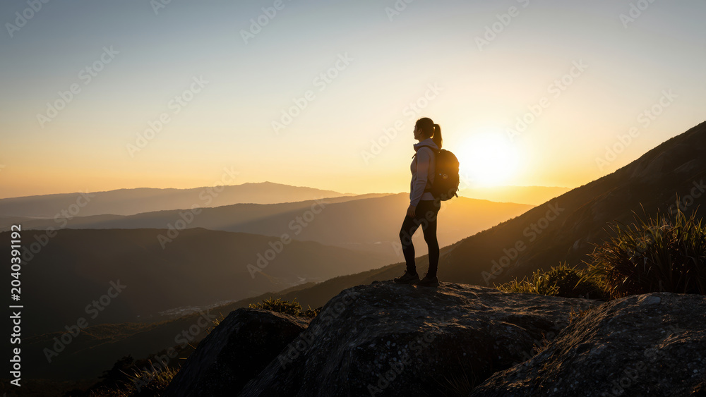 Obraz premium Summit Serenity: A lone hiker, silhouetted against a breathtaking sunset, stands on a rocky peak, gazing out at the vast, layered mountain landscape, embodying solitude and the spirit of adventure.