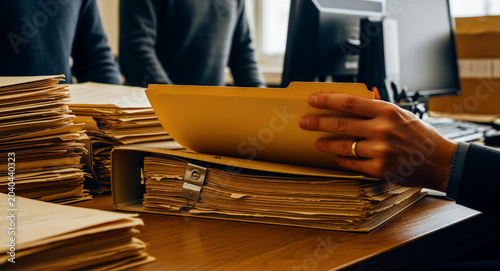 Hand opening a manila folder on a desk piled with documents and binders. Professional organizing extensive paperwork and archived files in a busy office. Document management and bureaucracy concept.