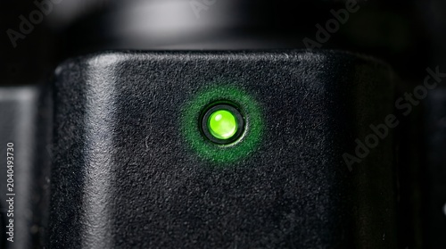 Glowing green LED status light on black plastic surface, electronic device power indicator lamp, macro shot of active signal light on hardware