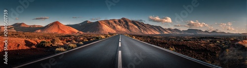 A long highway stretches into the distance, towards a mountain range under a blue, slightly cloudy sky