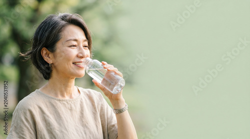 暑い日の水分補給を行う50代日本人女性の自然なライフスタイルシーン。
セージグリーン背景と自然光が爽やかな印象を演出。