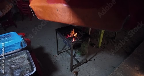 Close up of a small charcoal grill with glowing embers and flames under a makeshift orange canopy, with trays of fresh seafood nearby on a beach.