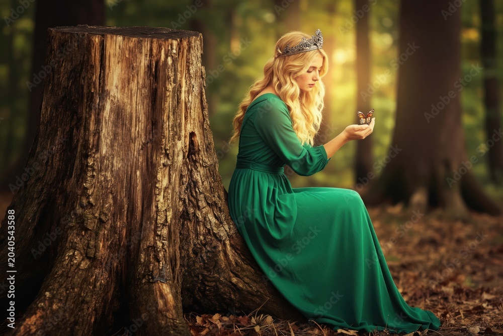 Obraz premium Young woman with blonde hair wears a green dress and crown. She sits by a tree stump in a sunlit forest holding a butterfly on her hand. Fantasy forest scene with a fairy tale mood.
