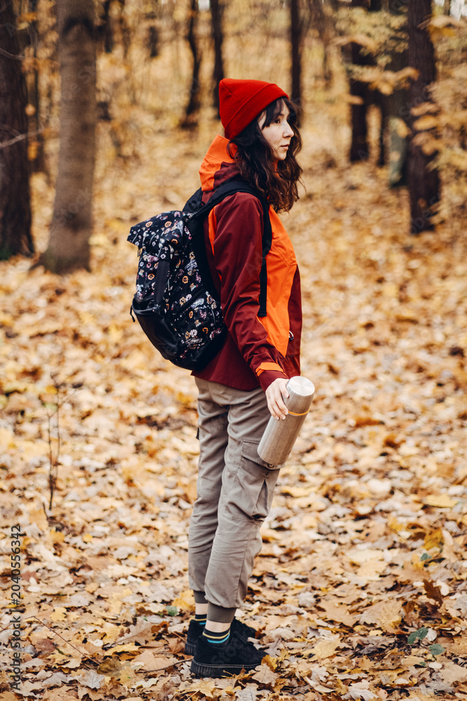 Naklejka premium Woman stands in autumn forest holding thermos. wilderness survival skills, outdoor escape plan, off-grid living preparation.