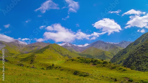 Time-lapse of white clouds flowing on blue sky above green mountains in Kyrgyzstan at sunny summer day. Cows grazing at the distance.