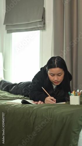 Young Asian Female Student Writing in Tablet on Bed in Cozy Dorm Room by Window