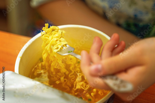 Teenage girl eating instant noodles from a cardboard box with a metal fork in her kitchen. Quick home meal moment