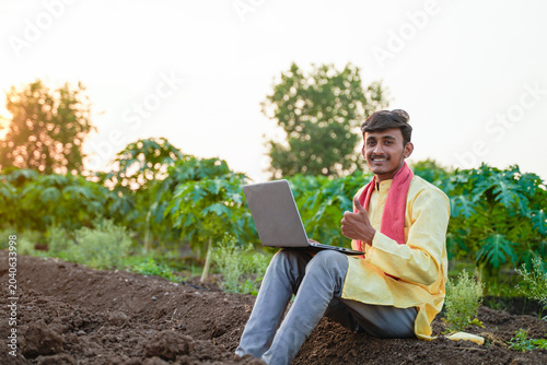 Obraz na płótnie Young Indian farmer using a laptop in an agricultural field, showcasing smart farming and digital agriculture at sunset