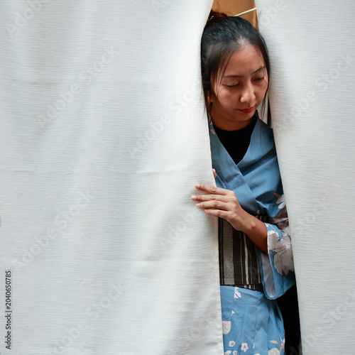 Japanese woman in blue floral kimono peeking through white noren curtain, Kyoto, Japan